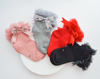 Op de afbeelding: Vier paar babysokken met kant en strikjes van satijnen lint. De sokken zijn roze, grijs, rood en zwart. De sokken zijn gemarkeerd met de maten S en M.