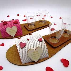 May include: Two wooden serving trays with clear plastic cups and decorative boxes. The boxes are pink and white with heart accents and the text "L'alcool è la migliore terapia di coppia". Red heart-shaped confetti is scattered around the trays.
