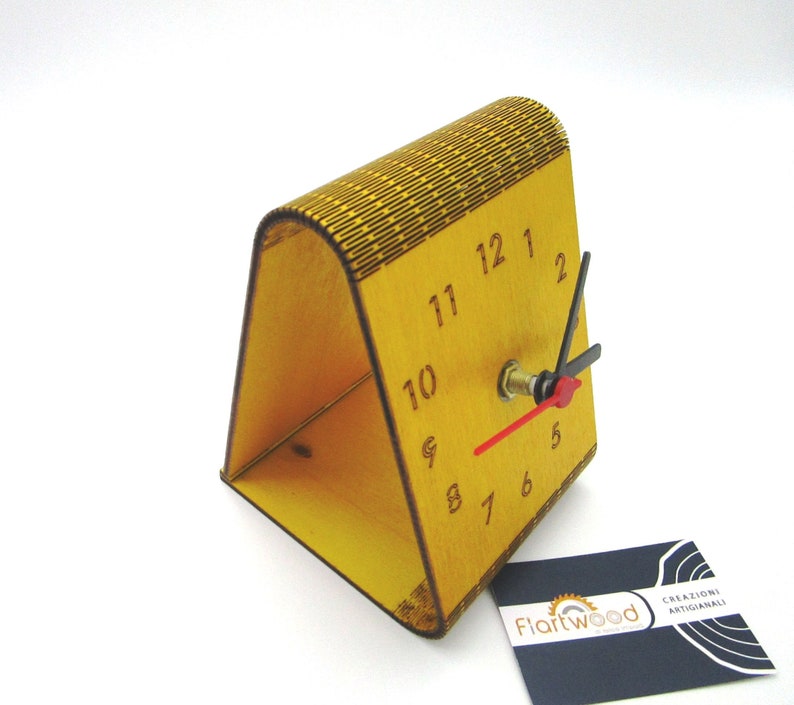 May include: A yellow wooden clock with black numbers and hands. The clock is on a stand and has a small business card with the text "Flartwood Creazioni Artigianali" in the bottom right corner.