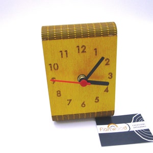 May include: A wooden clock with a yellow face and black hands. The clock is made of wood and has a decorative pattern on the front. The clock is sitting on a white surface. The clock is made by Fiartwood.