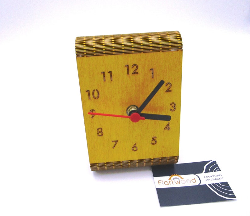 May include: A wooden clock with a yellow face and black hands. The clock is made of wood and has a decorative pattern on the front. The clock is sitting on a white surface. The clock is made by Fiartwood.