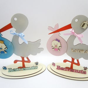 Może przedstawiać: Dwie drewniane figurki bocianów, jedna niebieska, a druga różowa, każda niosąca dziecko w torbie. Niebieski bocian ma napis "Benvenuto" na podstawie, a różowy bocian ma napis "Benvenuta" i imię "LAURA".