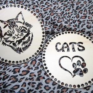 Könnte beinhalten: Zwei runde Holz-Stricknadeln mit Katzenmotiv. Eine Stricknadel zeigt eine Katze mit ausgeschnittenem Gesicht, die andere das Wort "CATS" mit einem ausgeschnittenen Herz und Pfotenabdruck.