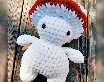 May include: White crocheted mushroom plush toy with a red and white crocheted hat. The toy is sitting on a wooden surface.