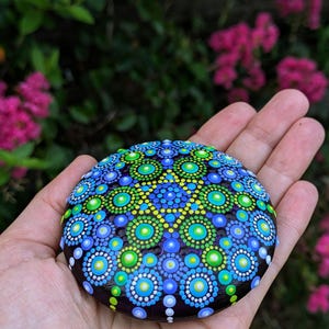May include: A hand holds a round, dark-colored stone painted with a colorful mandala design. The mandala features concentric circles of blue, green, and white dots.