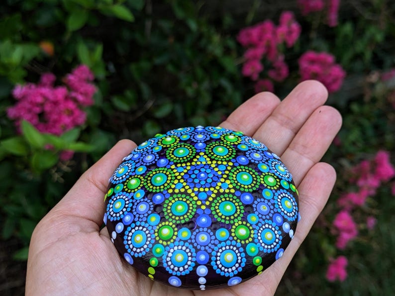 May include: A hand holds a round, dark-colored stone painted with a colorful mandala design. The mandala features concentric circles of blue, green, and white dots.