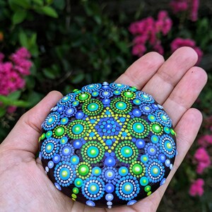 May include: A round, dark brown stone painted with a colorful mandala design. The mandala features concentric circles of blue, green, and yellow dots.