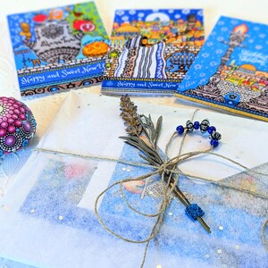 May include: A gift wrapped in blue and white tissue paper with a blue and white patterned ribbon. The gift is tied with a brown twine bow and decorated with a sprig of lavender and a string of blue beads. The gift is sitting on a white surface with three greeting cards in the background. The cards are decorated with images of Jerusalem and the text "Happy and Sweet New Year".