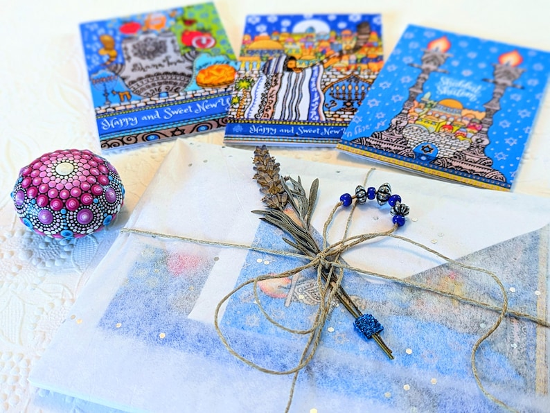 May include: A gift wrapped in blue and white tissue paper with a blue and white patterned ribbon. The gift is tied with a brown twine bow and decorated with a sprig of lavender and a string of blue beads. The gift is sitting on a white surface with three greeting cards in the background. The cards are decorated with images of Jerusalem and the text "Happy and Sweet New Year".
