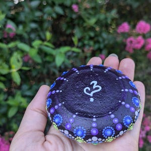 May include: A purple painted rock with a white heart and a question mark. The rock is decorated with small dots of purple, blue, green, and white paint.