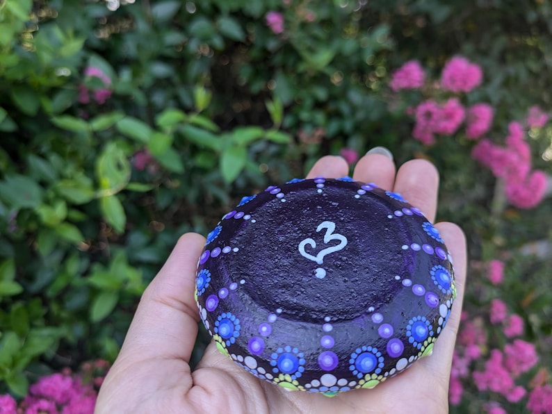 May include: A purple painted rock with a white heart and a question mark. The rock is decorated with small dots of purple, blue, green, and white paint.