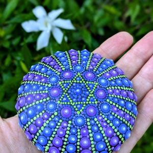 May include: A round, black stone painted with a mandala design in shades of blue, purple, and green. The mandala features a six-pointed star in the center.