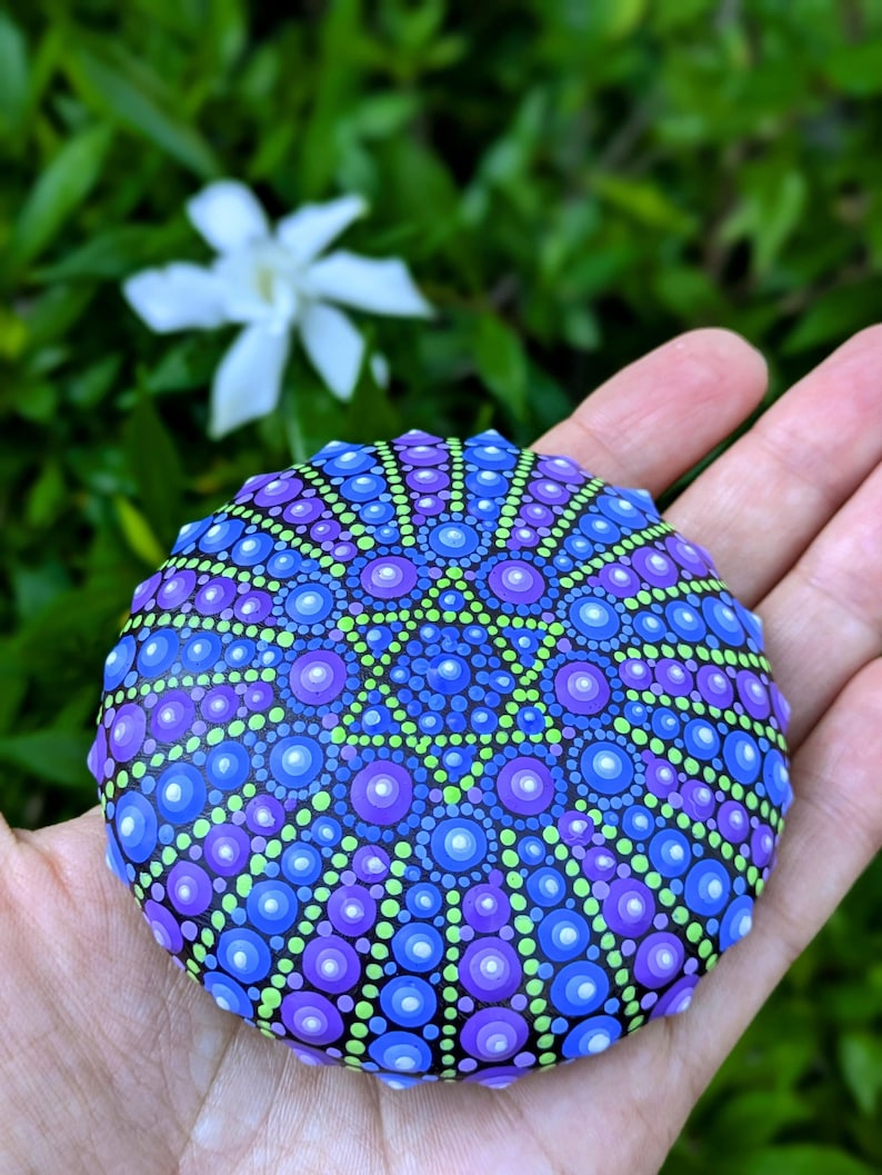 May include: A round, black stone painted with a mandala design in shades of blue, purple, and green. The mandala features a six-pointed star in the center.