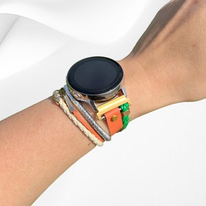 Puede incluir: Un smartwatch plateado con una esfera negra y una banda de cuero trenzado verde, naranja y blanco. El reloj se lleva en la muñeca.