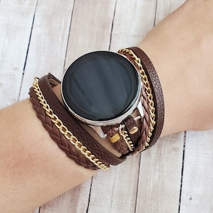 May include: Brown leather watch strap with gold chain detailing. The strap wraps around the wrist, creating a layered effect. The watch face is black and round, with a silver bezel. The strap is secured with a buckle.