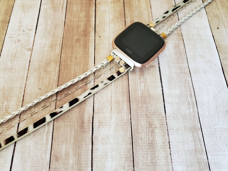 May include: A rose gold Fitbit smartwatch with a black screen is attached to a white braided strap, a silver chain, and a black and white animal print strap. The straps are attached to the watch with gold metal clasps.
