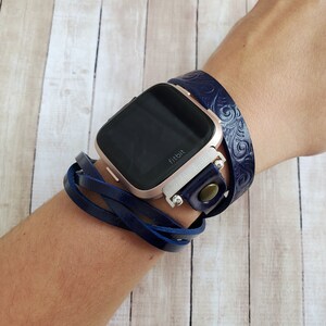 May include: A navy blue leather watch band with a decorative pattern wraps around a rose gold Fitbit smartwatch. The band is secured with a silver buckle and a brown button.