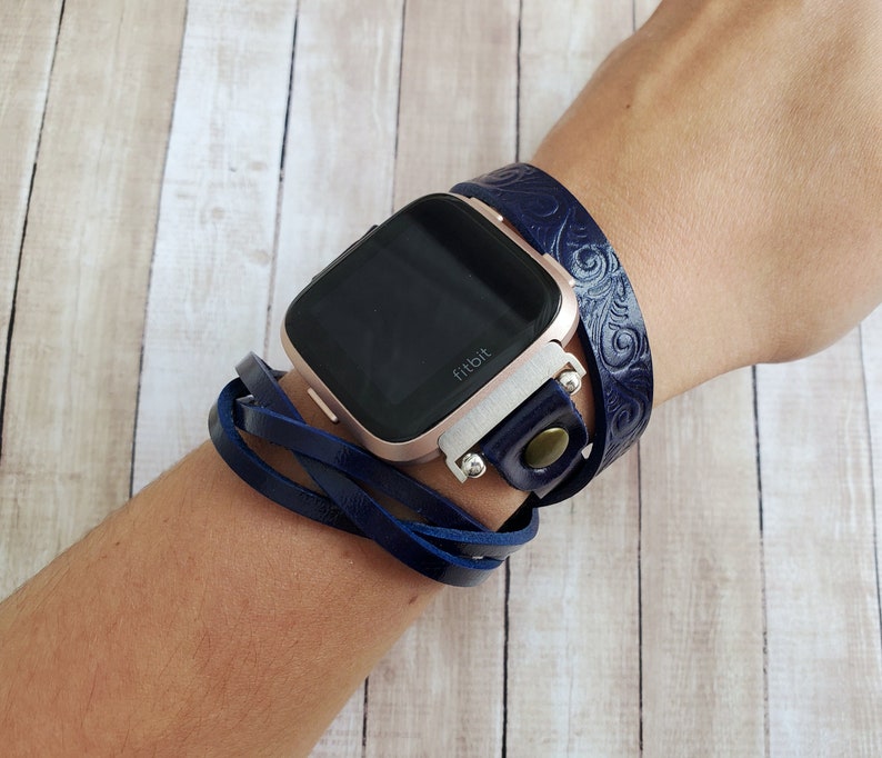 May include: A navy blue leather watch band with a decorative pattern wraps around a rose gold Fitbit smartwatch. The band is secured with a silver buckle and a brown button.