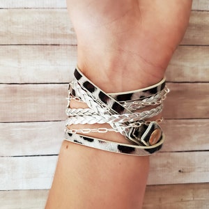 May include: A multi-layered bracelet with a white braided strap, a silver chain, and a leopard print strap. The bracelet has a gold button closure.