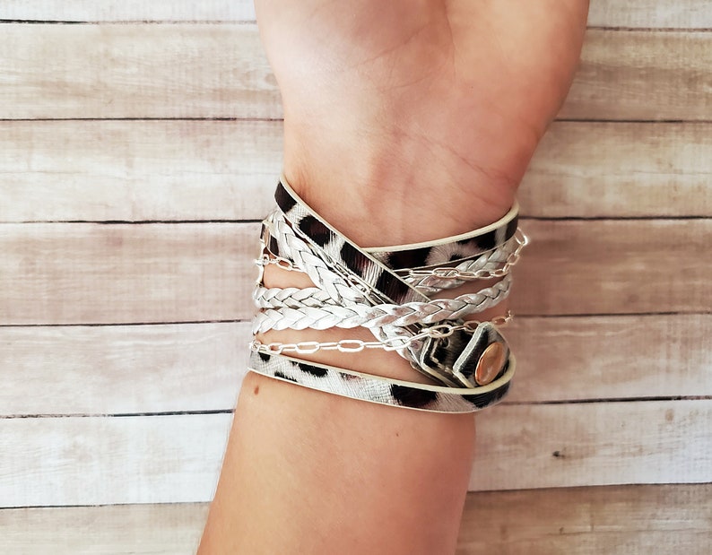 May include: A multi-layered bracelet with a white braided strap, a silver chain, and a leopard print strap. The bracelet has a gold button closure.