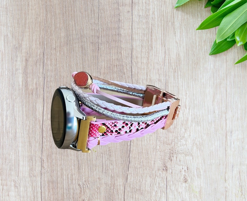 K&ouml;nnte beinhalten: Ein mehrstr&auml;ngiges Uhrenarmband in Rosa-, Wei&szlig;- und Silbert&ouml;nen mit einem ros&eacute;goldenen Verschluss. Das Armband hat geflochtene und gemusterte Elemente und ist an einer silbernen Smartwatch befestigt.