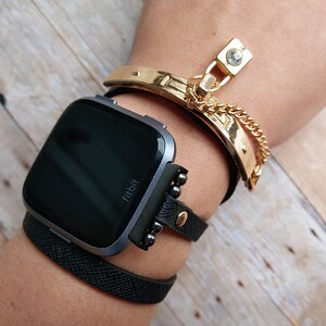 May include: A wrist adorned with a black Fitbit smartwatch and multiple bracelets. A gold-toned cuff bracelet with a chain and charm is paired with a black leather band and a textured black band. The watch has a dark screen.