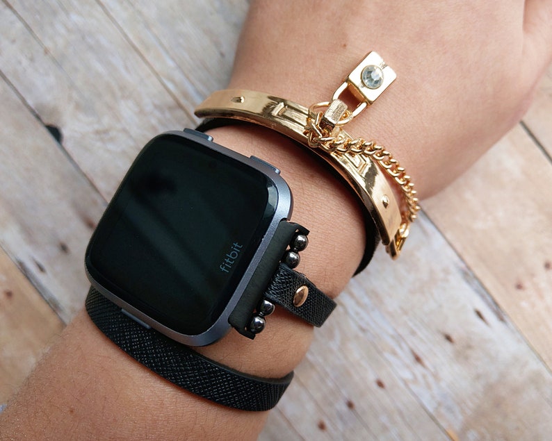 May include: A wrist adorned with a black Fitbit smartwatch and multiple bracelets. A gold-toned cuff bracelet with a chain and charm is paired with a black leather band and a textured black band. The watch has a dark screen.