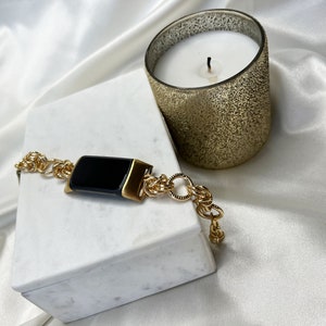 May include: A gold chain bracelet with a black rectangular watch face. The bracelet is on a white marble surface next to a gold candle.