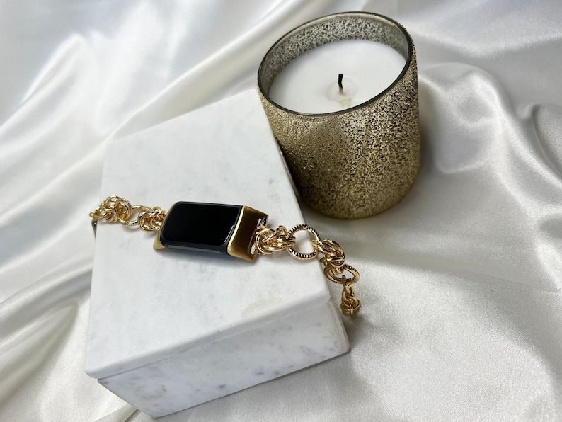 May include: A gold chain bracelet with a black rectangular watch face. The bracelet is on a white marble surface next to a gold candle.