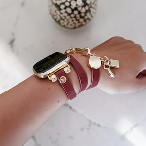 May include: A burgundy leather Apple Watch strap with gold-coloured hardware and decorative charms. The strap features a wrap-around design with a gold clasp and charms including a lock, key, and a round charm. The watch face is black and gold.