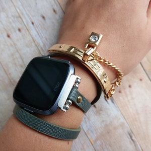 May include: A Fitbit Versa smartwatch with a dark screen is worn with a green leather band and a gold-colored bracelet. The gold bracelet has a decorative clasp and a chain. The watch band has silver-colored studs.