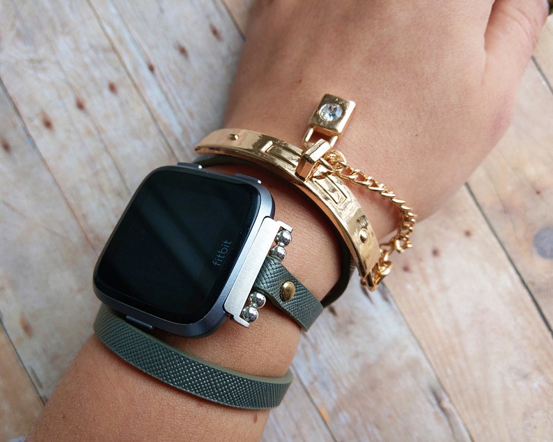 May include: A Fitbit Versa smartwatch with a dark screen is worn with a green leather band and a gold-colored bracelet. The gold bracelet has a decorative clasp and a chain. The watch band has silver-colored studs.