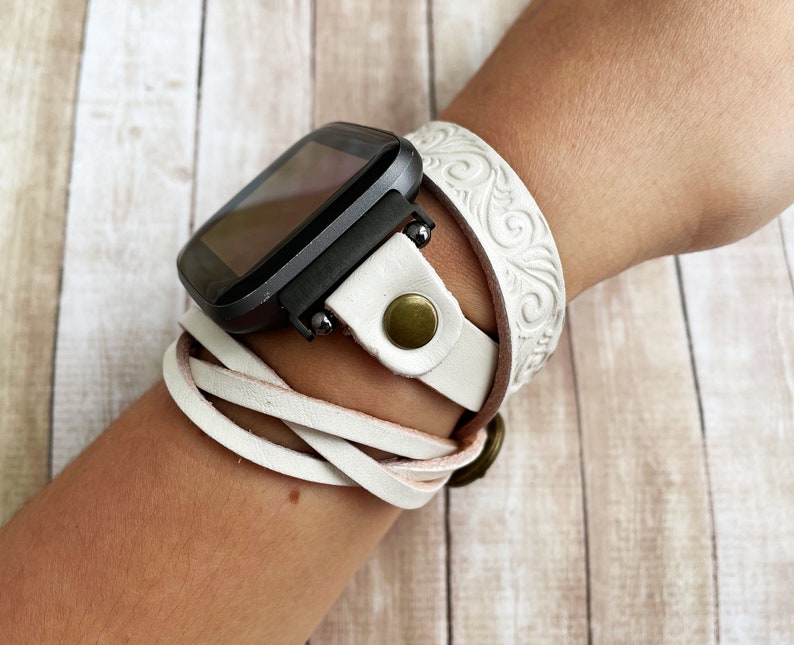 May include: White leather watch band with a decorative embossed design. The band is wrapped around a wrist and features a gold-toned buckle.