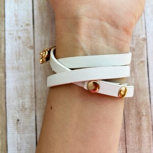May include: White wrap bracelet with gold-colored metal accents. The bracelet wraps around the wrist multiple times and fastens with a gold-colored clasp and two gold-colored snap closures. The bracelet is made of a textured material.