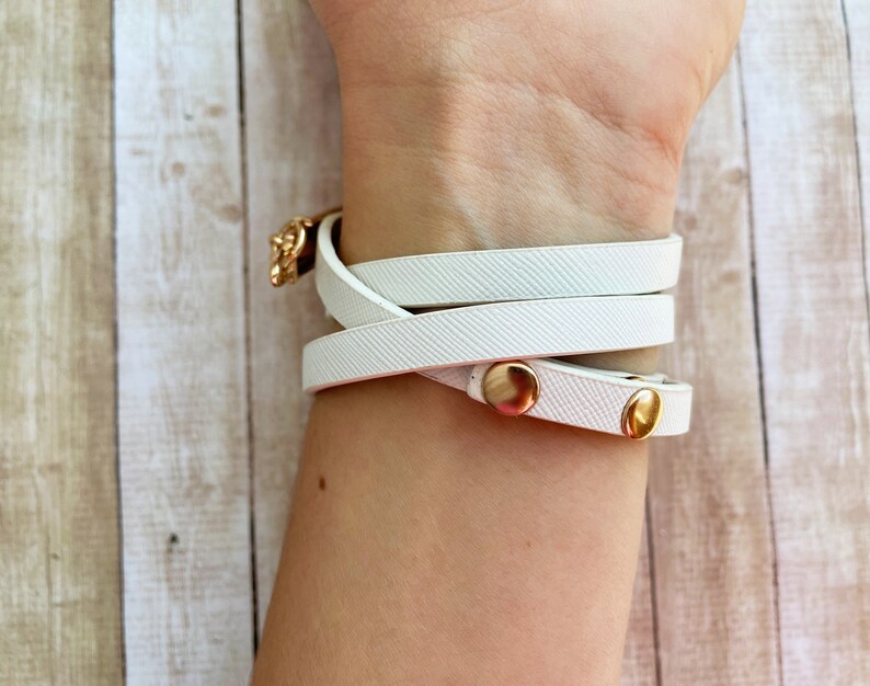 May include: White wrap bracelet with gold-colored metal accents. The bracelet wraps around the wrist multiple times and fastens with a gold-colored clasp and two gold-colored snap closures. The bracelet is made of a textured material.