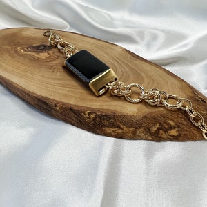 May include: A gold chain bracelet with a black rectangular watch face. The bracelet is on a wooden surface.