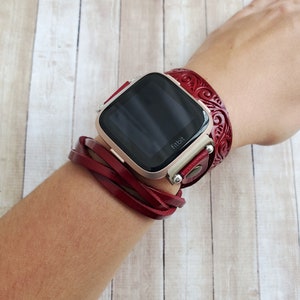 May include: A red leather watch band with a decorative design wraps around a wrist. The watch face is black and displays the word "fitbit".