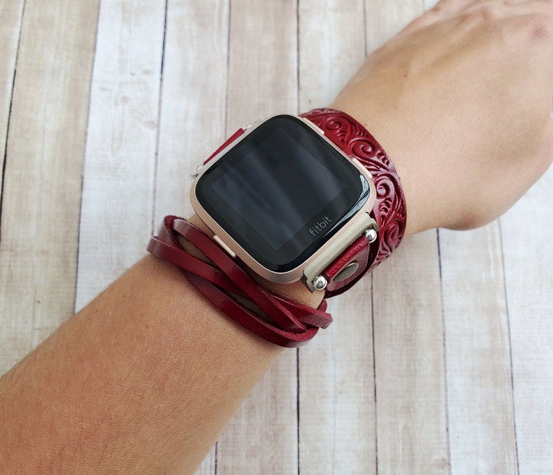 May include: A red leather watch band with a decorative design wraps around a wrist. The watch face is black and displays the word "fitbit".