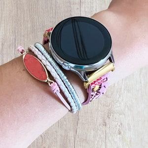 K&ouml;nnte beinhalten: Eine silberne Smartwatch mit schwarzem Bildschirm und einem Armband mit rosa Blumenmuster. Ein rosa-rotes Armband mit goldfarbenem Rahmen und ein geflochtenes wei&szlig;-silbernes Armband sind ebenfalls zu sehen.