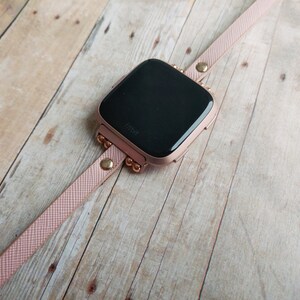 May include: A rose gold Fitbit smartwatch with a matching pink band. The watch face is black, and the band is secured with gold-colored studs and a buckle. The watch is displayed on a light-colored wooden surface.