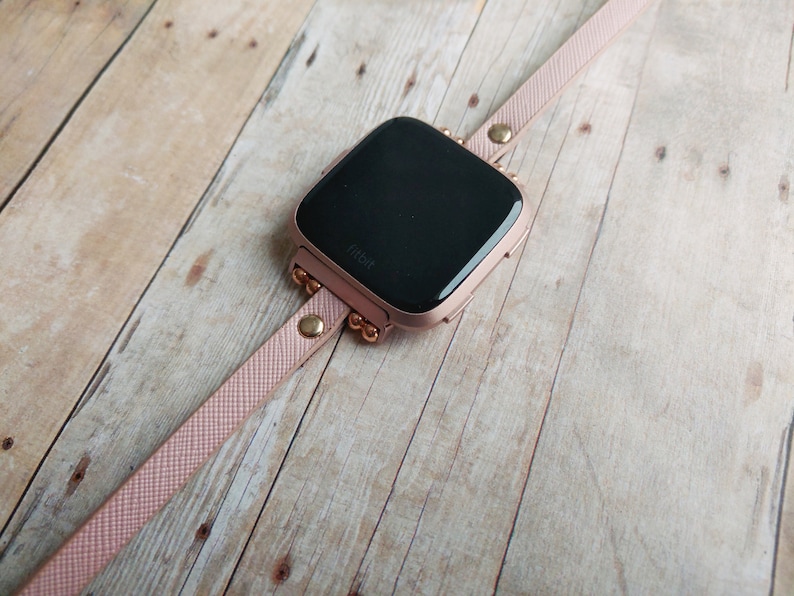 May include: A rose gold Fitbit smartwatch with a matching pink band. The watch face is black, and the band is secured with gold-colored studs and a buckle. The watch is displayed on a light-colored wooden surface.