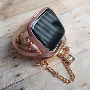 May include: A rose gold Fitbit smartwatch with a wrap-around leather band. The band is light pink and has gold-colored accents. A gold chain with a small lock charm hangs from the band. The watch is on a wooden surface.