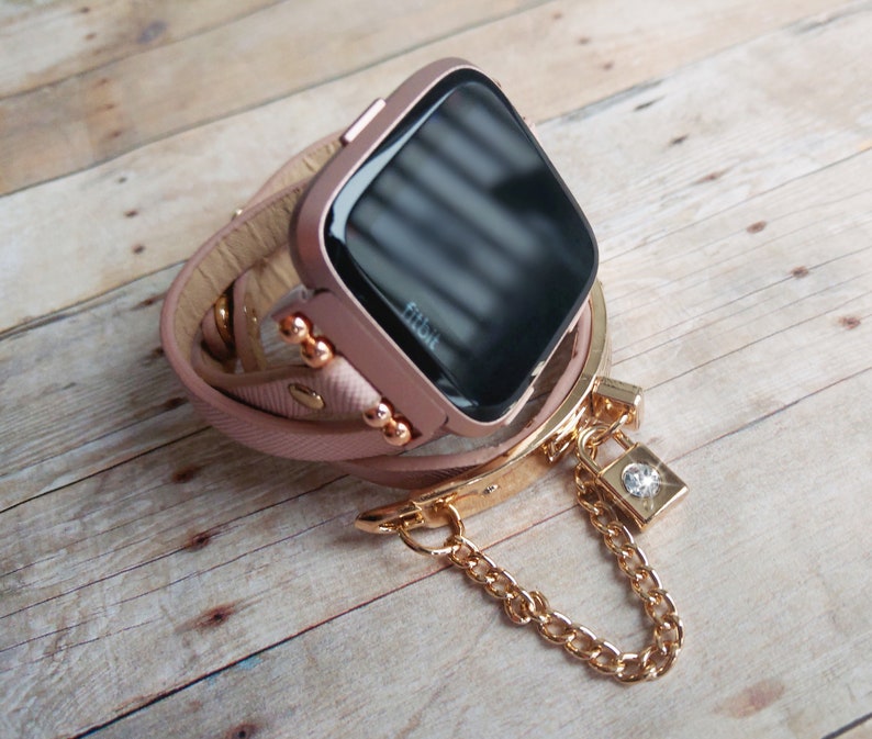 May include: A rose gold Fitbit smartwatch with a wrap-around leather band. The band is light pink and has gold-colored accents. A gold chain with a small lock charm hangs from the band. The watch is on a wooden surface.