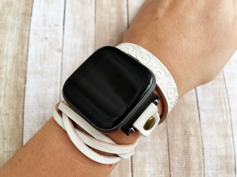 May include: White leather watch band with a decorative pattern and a black smartwatch. The band is wrapped around the wrist.
