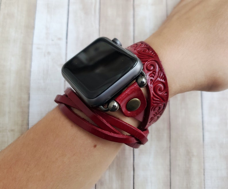 Może przedstawiać: Czerwony sk&oacute;rzany pasek do zegarka z ozdobnym wzorem. Pasek jest owinięty wok&oacute;ł nadgarstka i ma przymocowany srebrny zegarek Apple Watch.