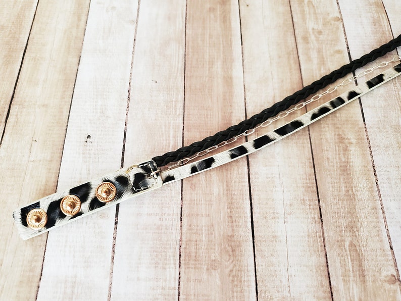 May include: A black braided cord, a silver chain, and a white and black leopard print leather strap are woven together to create a unique bracelet. The bracelet has gold-toned metal accents.