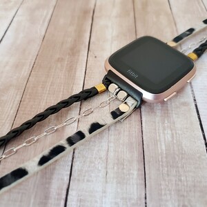 May include: A rose gold Fitbit smartwatch with a black band and a black braided strap, a silver chain, and a white and black animal print strap.