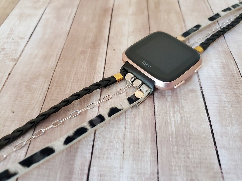 May include: A rose gold Fitbit smartwatch with a black band and a black braided strap, a silver chain, and a white and black animal print strap.
