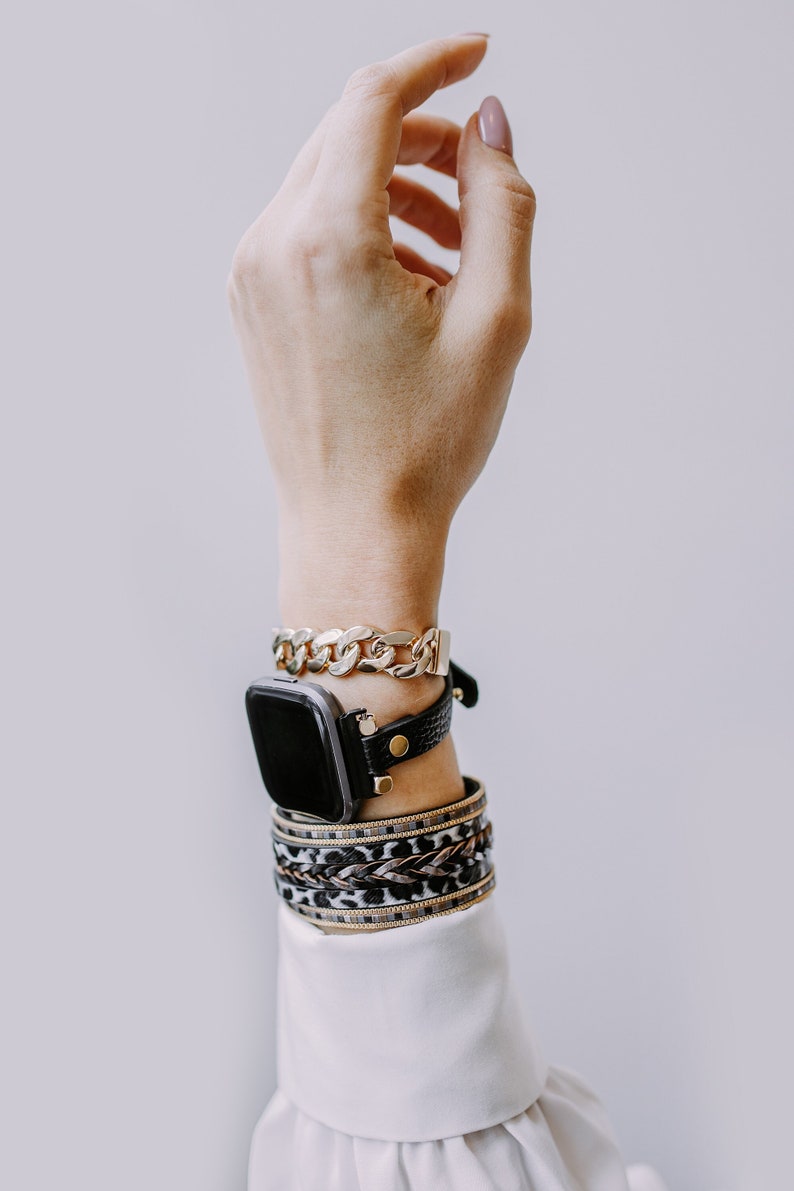 Peut inclure: Un poignet de femme avec un bracelet de montre en cuir noir et un bracelet en cha&icirc;ne dor&eacute;e. Le cadran de la montre est noir et carr&eacute;. La femme porte plusieurs bracelets au poignet, dont un bracelet &agrave; imprim&eacute; l&eacute;opard et un bracelet en or.