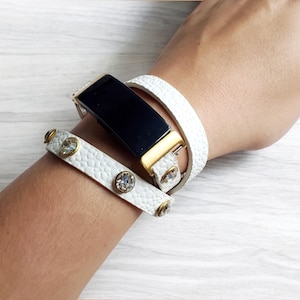 Puede incluir: Una colección de pulseras de cuero blanco y un reloj inteligente negro con detalles dorados. Una pulsera presenta piedras preciosas redondas y transparentes y tachuelas doradas. Las pulseras están apiladas en una muñeca, mostrando un estilo de accesorio moderno.